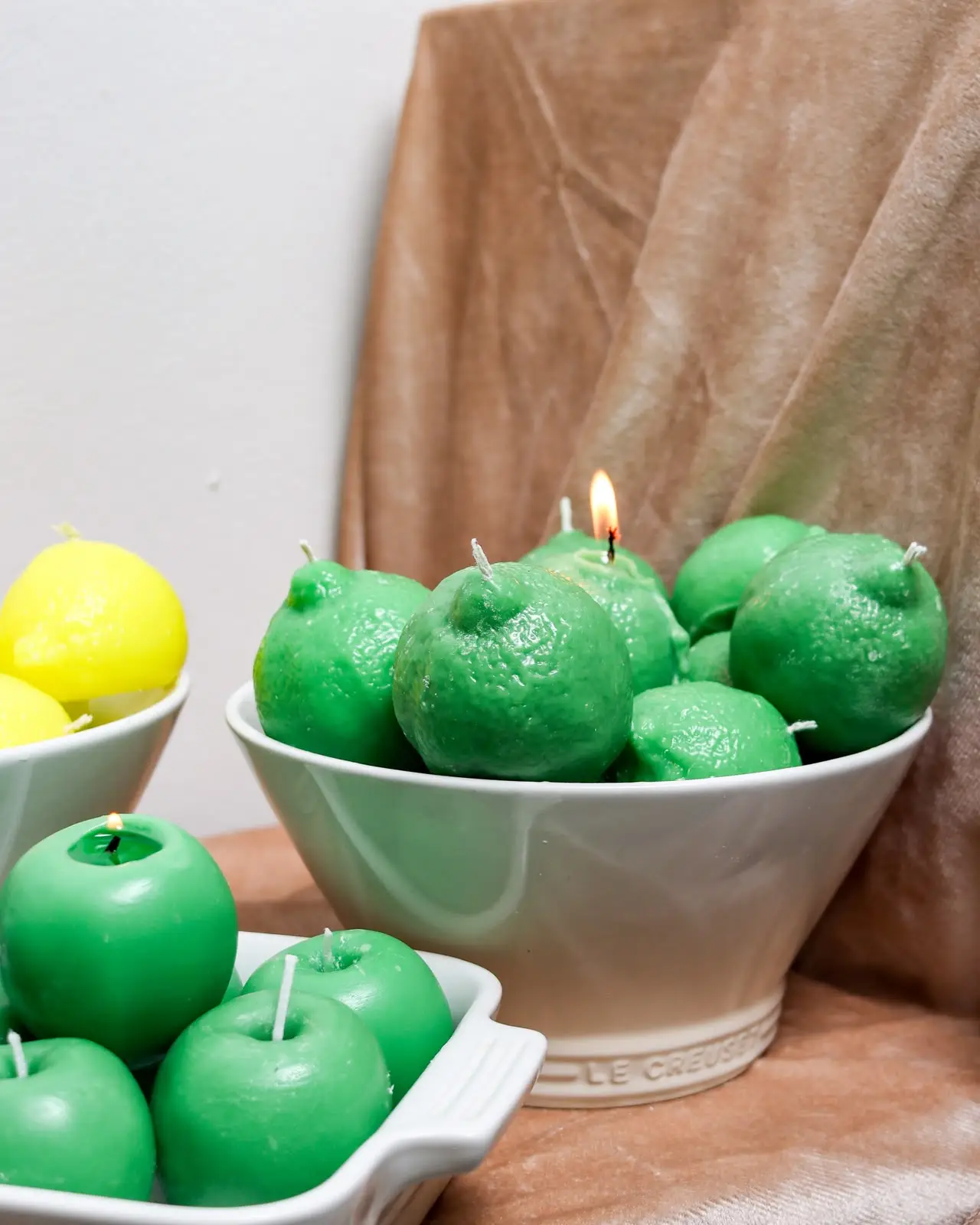 Green lemon candles in a big white bowl with apples and more lemons around it on a stand.